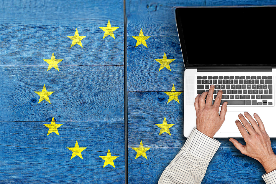 Person typing on a laptop keyboard on a blue wooden table, surrounded by yellow stars from the EU flag.
