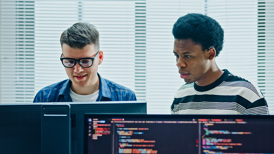 Two people sitting at monitors, collaboratively working on computer code in a modern office.