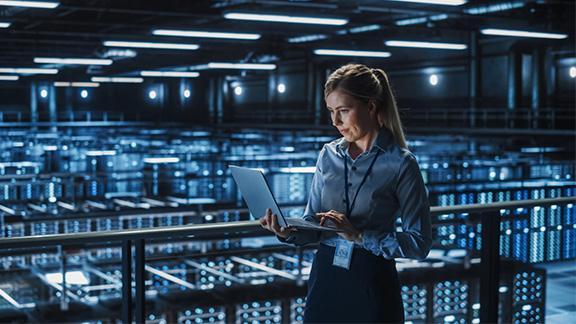 A woman stands in a server room with an open laptop in her hands